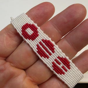 Puede incluir: Pulsera de amistad blanca y roja hecha a mano. La pulsera presenta un patr&oacute;n repetitivo de formas geom&eacute;tricas rojas sobre un fondo blanco. La pulsera est&aacute; hecha de hilos tejidos.