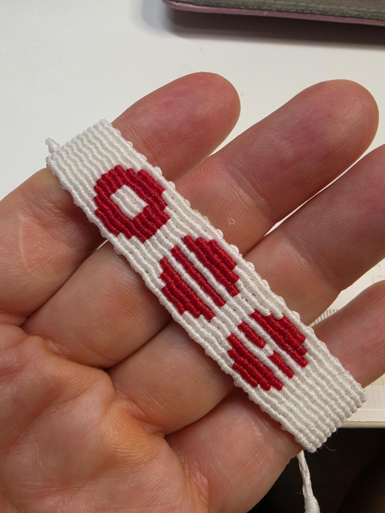 Puede incluir: Pulsera de amistad blanca y roja hecha a mano. La pulsera presenta un patr&oacute;n repetitivo de formas geom&eacute;tricas rojas sobre un fondo blanco. La pulsera est&aacute; hecha de hilos tejidos.