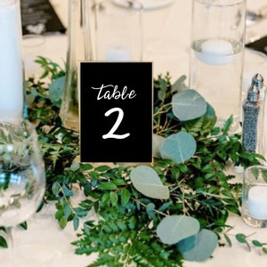 May include: A black table number card with gold trim and white text that reads "Table 2". The card is surrounded by greenery.
