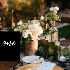 May include: A black card with the word "one" written in white cursive script. The card is on a wooden table with white flowers and greenery in vases.