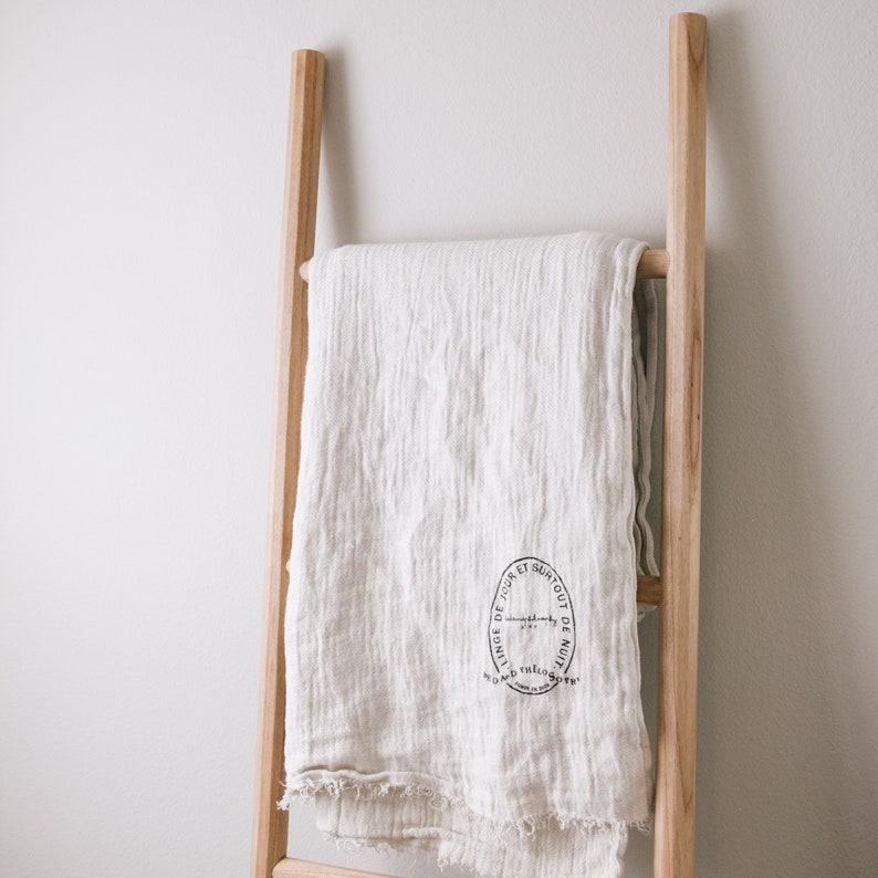 Puede incluir: Una manta de lino blanca con un sello negro que dice "Linge de Jour et Surtout de Nuit" drapeada sobre una escalera de madera.