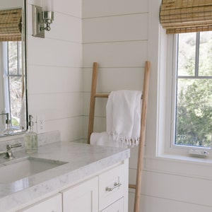 Puede incluir: Un ba&ntilde;o blanco con una escalera de madera apoyada en la pared. Una toalla blanca est&aacute; colgada sobre la escalera. El ba&ntilde;o tiene un tocador blanco con una encimera de m&aacute;rmol y un lavabo blanco. Hay una ventana con persianas de bamb&uacute; en el fondo.