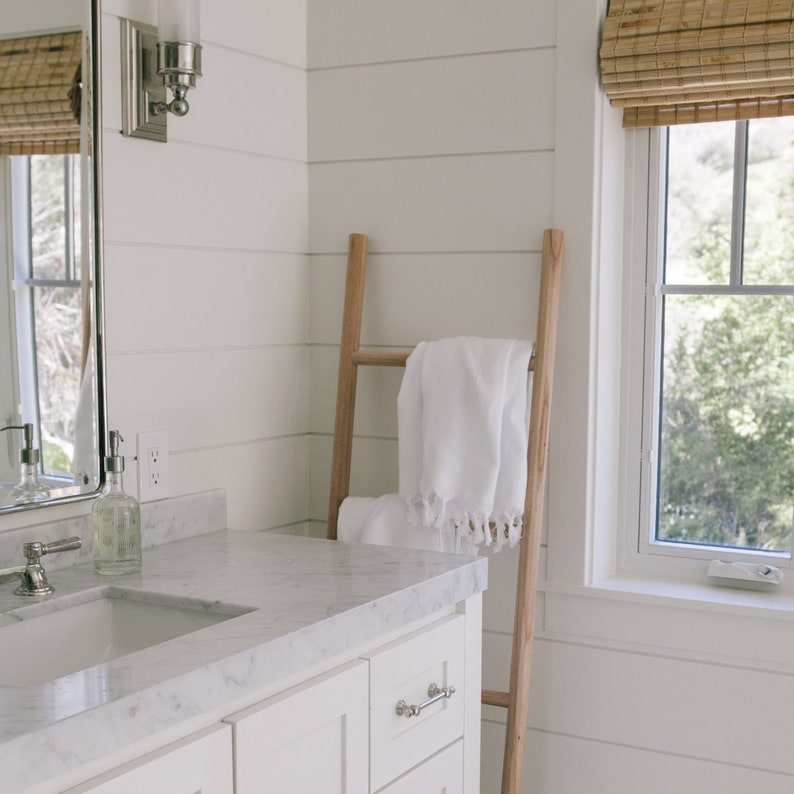 Puede incluir: Un ba&ntilde;o blanco con una escalera de madera apoyada en la pared. Una toalla blanca est&aacute; colgada sobre la escalera. El ba&ntilde;o tiene un tocador blanco con una encimera de m&aacute;rmol y un lavabo blanco. Hay una ventana con persianas de bamb&uacute; en el fondo.