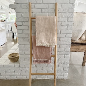 Puede incluir: Una escalera de madera apoyada en una pared de ladrillo blanco con dos mantas rosas y blancas colgadas sobre ella.
