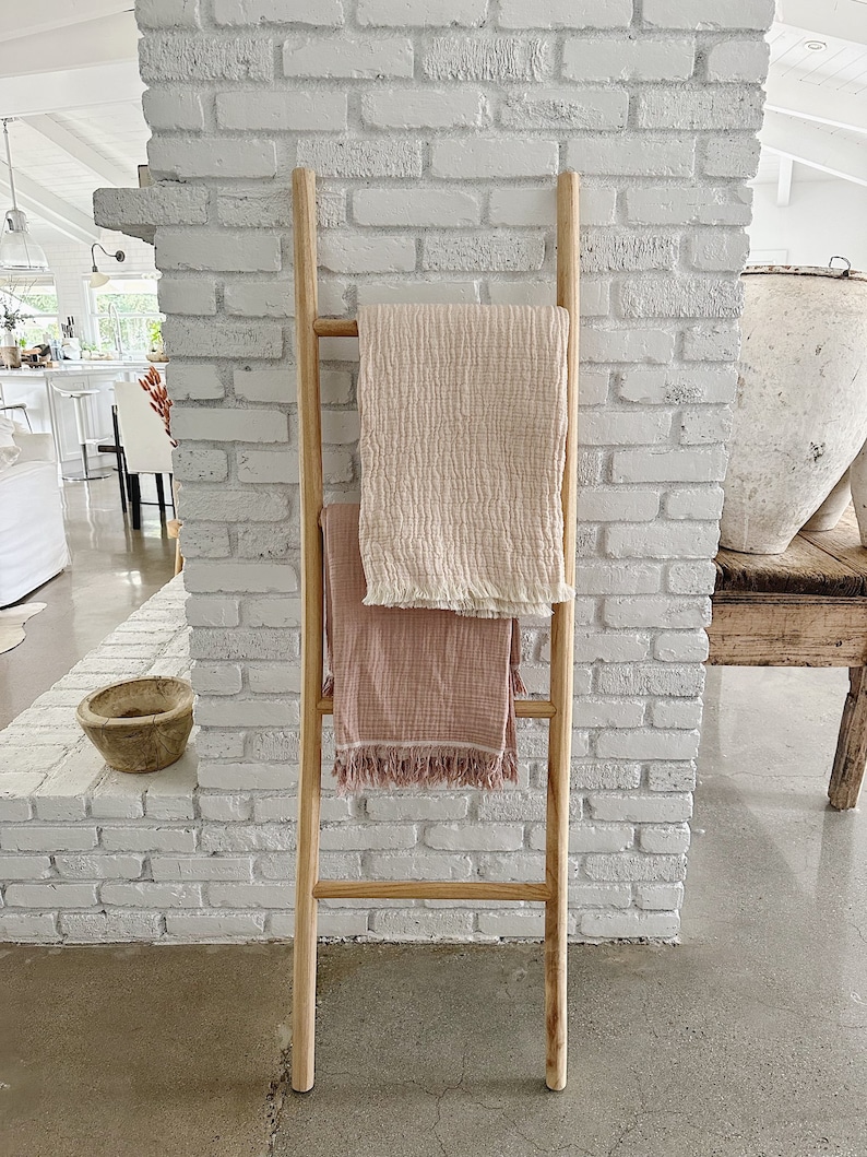 Puede incluir: Una escalera de madera apoyada en una pared de ladrillo blanco con dos mantas rosas y blancas colgadas sobre ella.