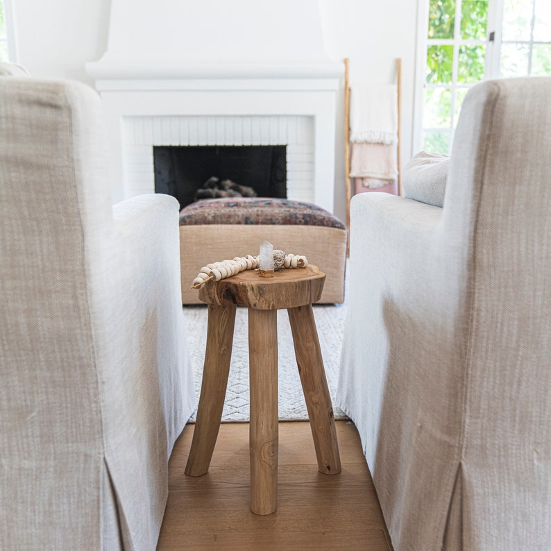 beautiful handmade teak stool in between two restoration hardware love seats with a handmade blanket ladder leaning against a white wall next to a fireplace