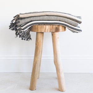 May include: A wooden stool with four legs and a natural wood finish. A black and white striped throw blanket is draped over the stool.