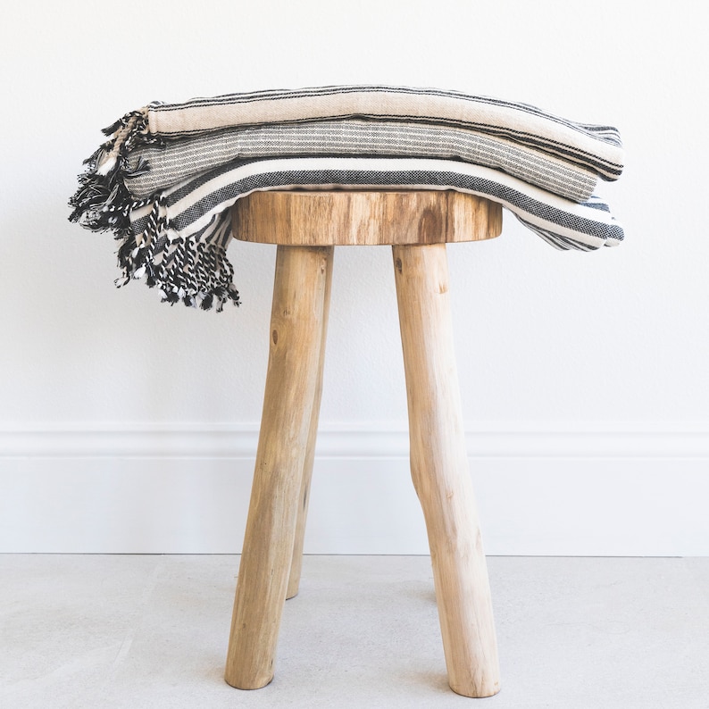 May include: A wooden stool with four legs and a natural wood finish. A black and white striped throw blanket is draped over the stool.
