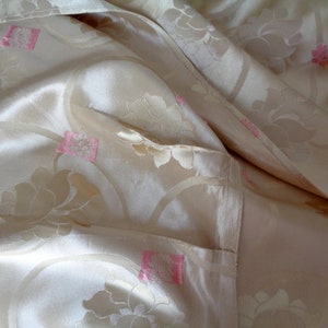 May include: A close-up of a cream-colored fabric with a floral pattern. The fabric has a subtle sheen and is decorated with pink flowers.