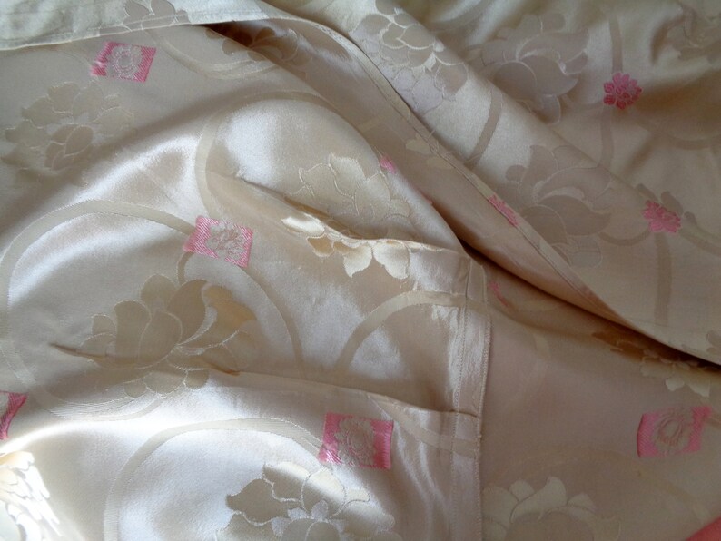 May include: A close-up of a cream-colored fabric with a floral pattern. The fabric has a subtle sheen and is decorated with pink flowers.