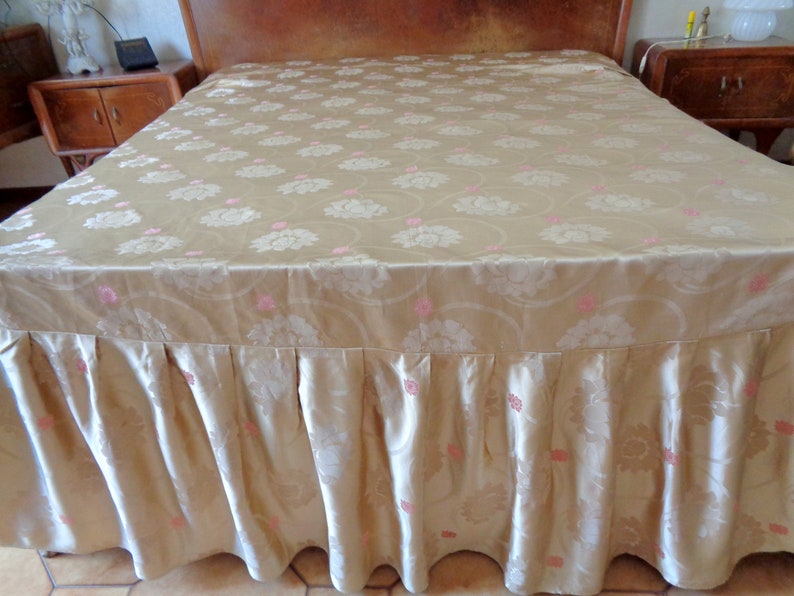 May include: A gold bed skirt with a pink floral pattern. The skirt has a ruffled edge and is made of a silky fabric.