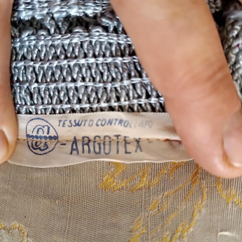 May include: A close-up of a fabric label with the text "TESSUTO CONTROLLATO" and "-ARGOTEX-". The label is cream-coloured with blue text and a circular logo. The fabric beneath is beige with gold embroidery, and the top fabric is silver and grey.