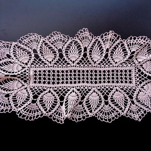 May include: A delicate, crocheted table runner in a light pink color. The runner features a repeating pattern of intricate lacework with a central rectangular section.