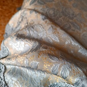 May include: Close-up of a folded fabric with a floral pattern. The fabric features silver, gold, and cream colours. The design includes detailed floral and leaf motifs, creating a textured appearance. The edges of the fabric are trimmed with a silver fringe.