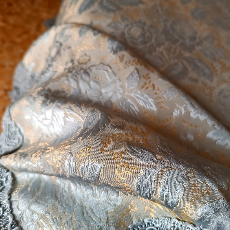 May include: Close-up of a folded fabric with a floral pattern. The fabric features silver, gold, and cream colours. The design includes detailed floral and leaf motifs, creating a textured appearance. The edges of the fabric are trimmed with a silver fringe.