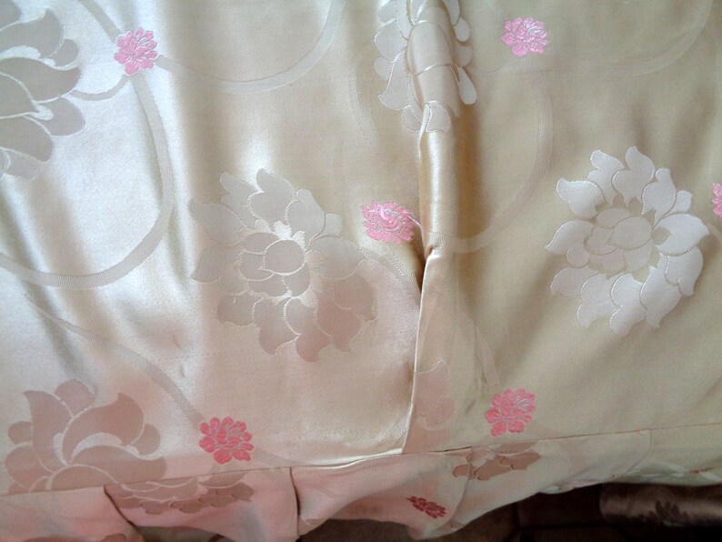 May include: A close-up of a cream-colored fabric with a floral pattern. The fabric has a subtle sheen and is decorated with pink flowers.