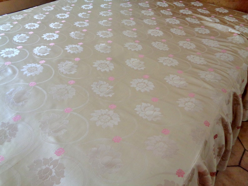May include: A white bedspread with a delicate floral pattern in pink. The bedspread has a ruffled edge.