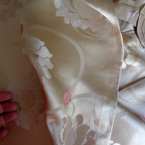 May include: A close-up of a cream-colored fabric with a floral pattern. The fabric has a subtle sheen and a delicate texture. The flowers are embossed and have a pink hue.