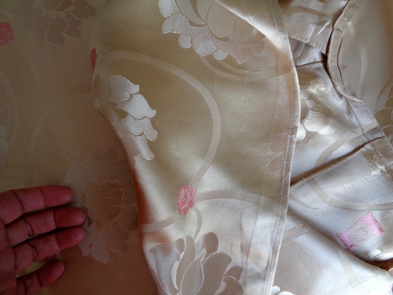 May include: A close-up of a cream-colored fabric with a floral pattern. The fabric has a subtle sheen and a delicate texture. The flowers are embossed and have a pink hue.