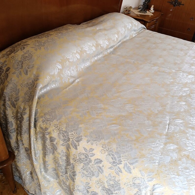 May include: A bed with a floral patterned bedspread. The bedspread has a repeating pattern of grey and silver flowers on a cream background. The bedspread is draped over the bed, with the wooden headboard visible in the background.