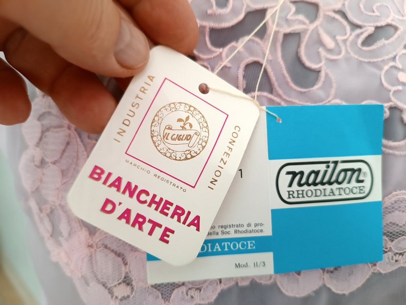 May include: A white tag with the text "INDUSTRIA BIANCHERIA D'ARTE" and a logo of a flower with the text "IL GIGLIO" and "MARCHIO REGISTRATO". A blue tag with the text "nailon RHODIATOCE" and "Mod. 11/3".