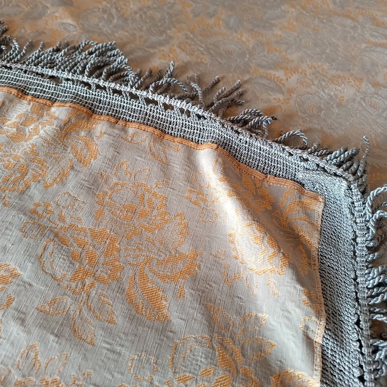 May include: A close-up shot of a decorative textile featuring a floral pattern in orange and grey. The fabric has a grey crocheted border with fringe. This could be a pillow or a decorative item for the home.