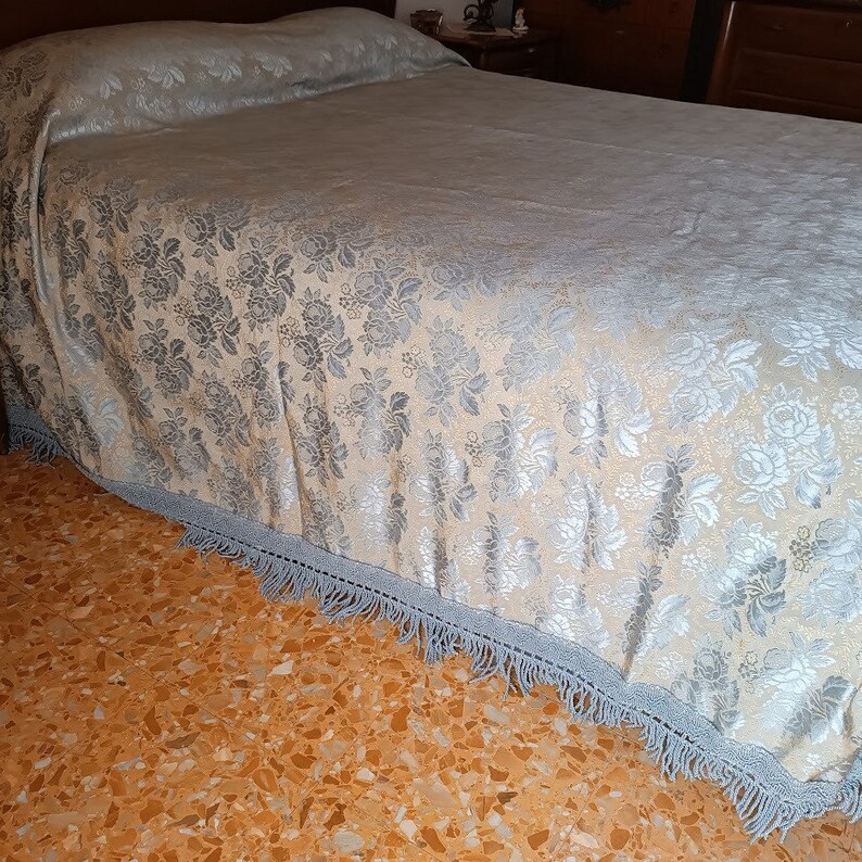 May include: A bedspread with a floral pattern in silver and cream. The bedspread has a light blue fringe trim. The bedspread is on a bed with a wooden headboard.