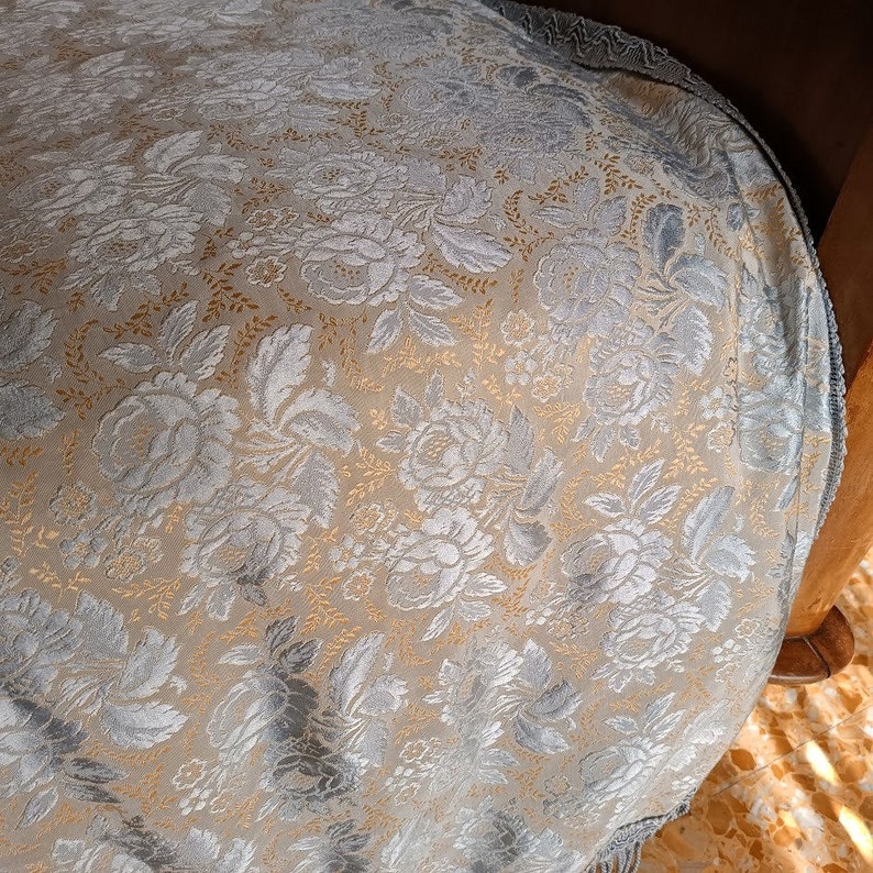 May include: Close-up of a floral patterned fabric in shades of silver, gold, and beige. The design features detailed roses and leaves. The fabric is draped over a wooden chair.