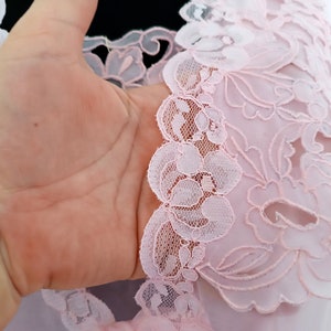 May include: Pink lace fabric with a floral design. The lace is delicate and intricate, with a scalloped edge.