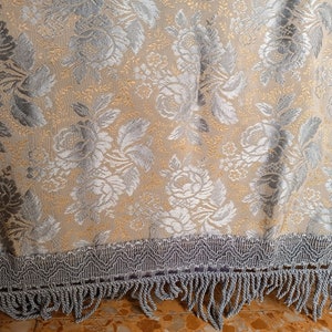 May include: A close-up of a beige floral fabric with silver and gold accents. The fabric features a repeating pattern of roses and leaves. The bottom edge has a silver fringe trim.