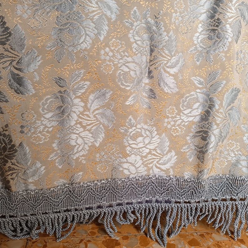 May include: A close-up of a beige floral fabric with silver and gold accents. The fabric features a repeating pattern of roses and leaves. The bottom edge has a silver fringe trim.
