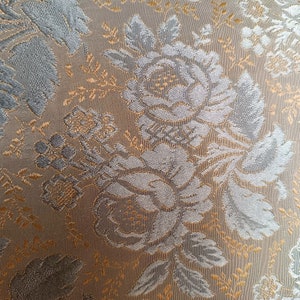May include: Close-up of a fabric with a floral pattern. The design features large, stylized roses and leaves in shades of grey and silver, set against a tan and gold background. The fabric has a textured appearance.