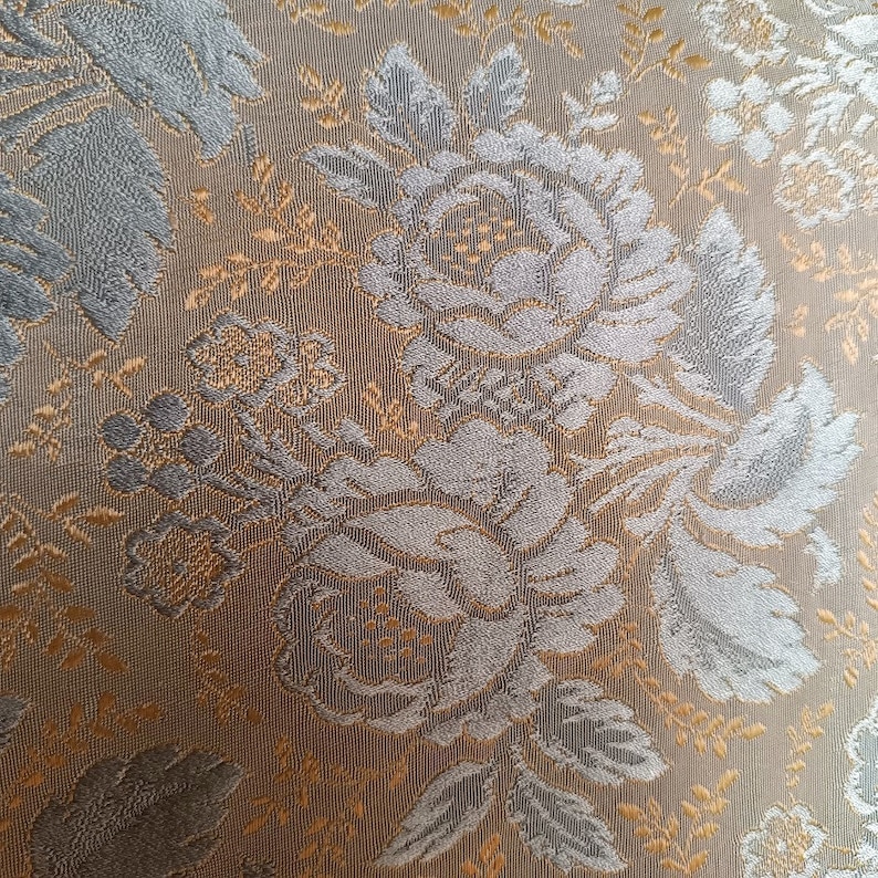 May include: Close-up of a fabric with a floral pattern. The design features large, stylized roses and leaves in shades of grey and silver, set against a tan and gold background. The fabric has a textured appearance.