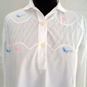 Può includere: Una camicia bianca a maniche lunghe con colletto e chiusura con bottoni. La camicia presenta un motivo a rombi ricamato con filo rosa e blu. Il ricamo include anche disegni floreali in rosa e blu.
