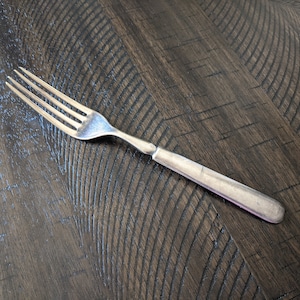 May include: A silver fork with four prongs, lying on a dark brown wooden surface. The fork has a simple, elegant design, with a smooth handle and a polished finish. The wooden surface has a textured, grooved pattern.