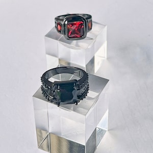 May include: Two black rings with square-shaped gemstones. The top ring has a red gemstone, and the bottom ring has a black gemstone.