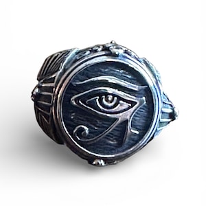 May include: A silver-toned ring featuring the Eye of Horus symbol. The ring has a circular face with the eye design in the center, surrounded by ornate detailing. The ring's band has a textured appearance, and the overall design is detailed and intricate.
