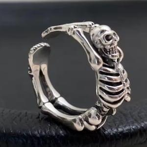 May include: A silver-colored ring shaped like a skeleton. The ring features a detailed skull and rib cage design, with articulated arms and legs. The ring is shown on a dark surface.