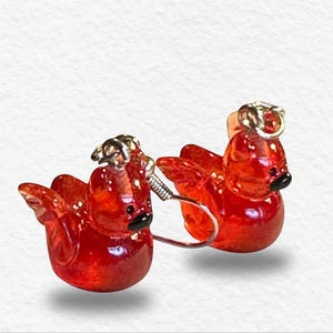 May include: A pair of red bird-shaped earrings with silver-coloured ear wires. The birds have black beaks and small wings. The earrings are made of a translucent material, and the birds have a glossy finish.