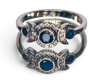 May include: Two silver rings stacked on top of each other. Each ring features a central dark blue gemstone surrounded by smaller clear stones. Crescent moon shapes flank the center stone, with small blue gems on each side.