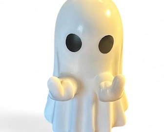 May include: A white ghost ornament with black eyes and arms extended in a welcoming gesture. The ghost has a rounded head and a draped body. The figurine is likely made of ceramic or a similar material.