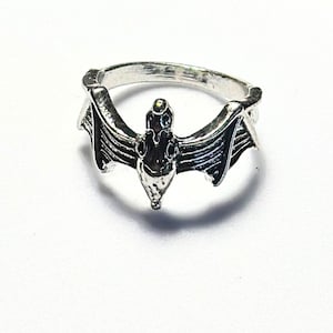 May include: A silver ring with a bat design. The bat's wings are spread out and the body is detailed with lines and curves.