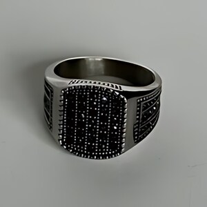 May include: A silver ring with a black square-shaped stone set in the center. The stone is surrounded by a silver border and the ring has a textured band.