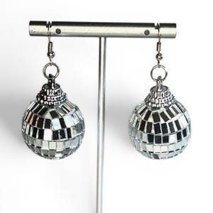 Puede incluir: Un par de pendientes de bola de discoteca plateados. Cada pendiente presenta un pequeño diseño de bola de discoteca redonda con azulejos cuadrados reflectantes. Los pendientes cuelgan de alambres de gancho plateados, creando un accesorio divertido.