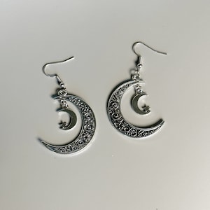 May include: A pair of silver earrings featuring a crescent moon design with intricate details and a small star charm hanging from the bottom.