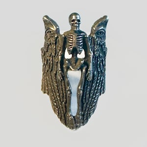 Może przedstawiać: Srebrny pierścionek z figurą szkieletu stojącą między dwiema postaciami z skrzydłami. Skrzydła są ozdobione piórami, a szkielet ma czaszkę.
