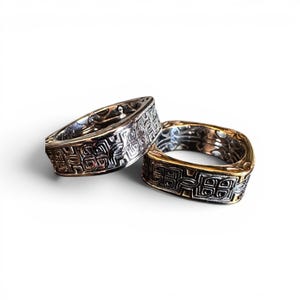 May include: Two square rings with detailed, repeating patterns. One ring is silver-toned, the other has a gold-toned band. The rings are probably metal and are shown against a white backdrop.