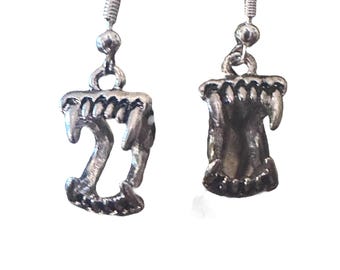 May include: A pair of silver-colored earrings. Each earring features a detailed charm shaped like a set of vampire fangs. The fangs are attached to a simple hook for pierced ears. The earrings are set against a white background.