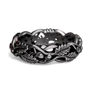 Puede incluir: Un anillo gris oscuro con un intrincado diseño de hojas de roble y bellotas. El anillo es de metal y tiene una superficie detallada y texturizada. El diseño del anillo tiene un tema inspirado en la naturaleza.