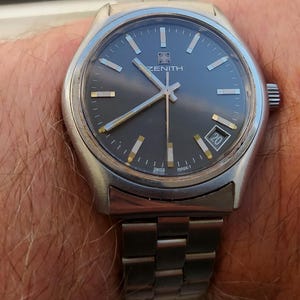 Pode incluir: Um relógio de pulso Zenith prateado com mostrador cinza escuro e visor de data às 3 horas. O relógio tem ponteiros e marcadores prateados e uma pulseira de metal prateado. O relógio está marcado "SWISS MADE".